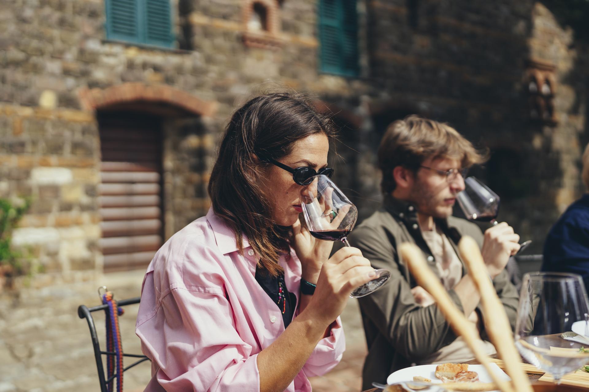 Guests nosing a glass of red wine at a tasting table outside a Tuscan stone farmhouse