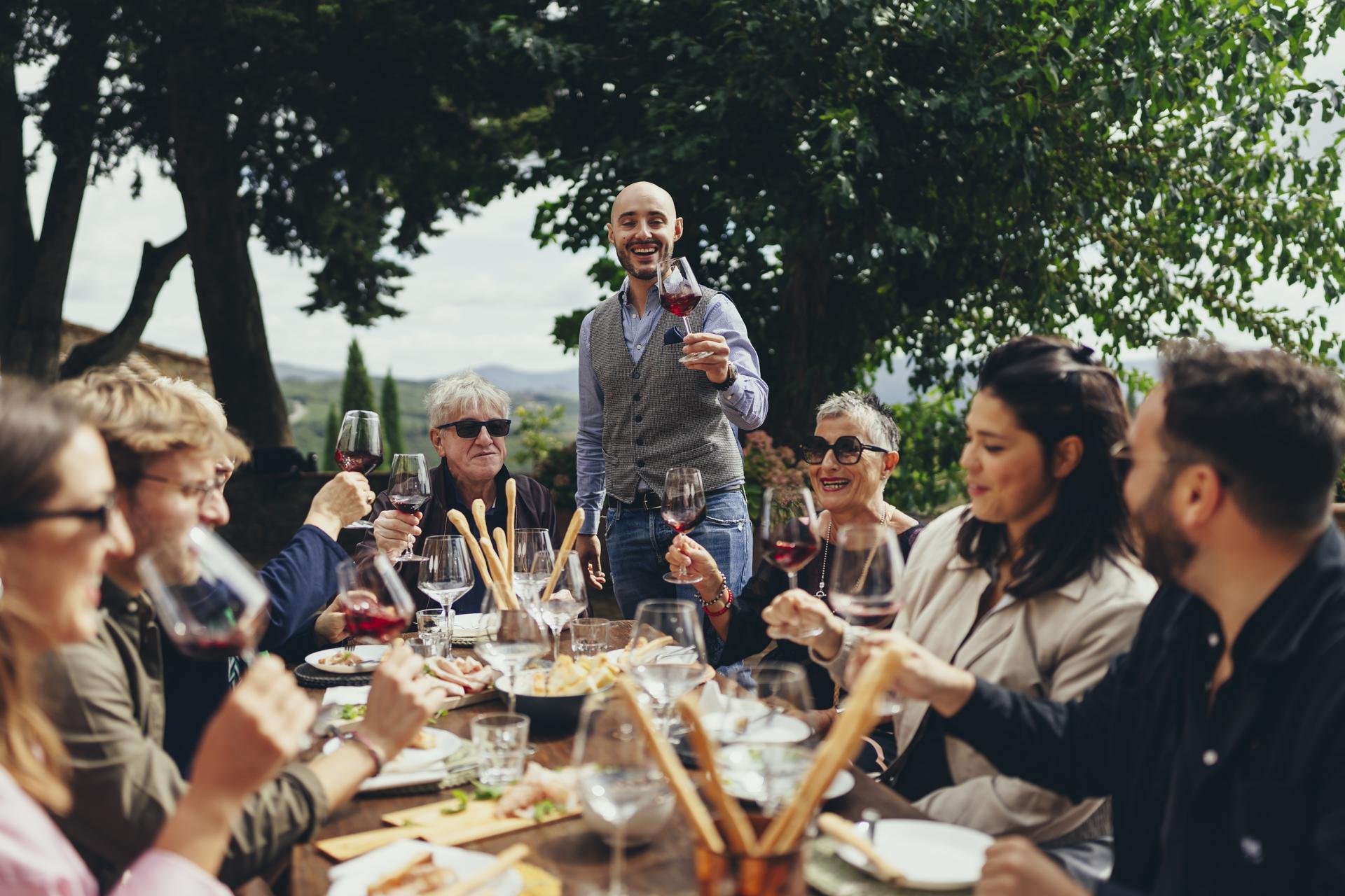 A sommelier raising a glass of red wine with guests at an outdoor tasting lunch in the Tuscan hills