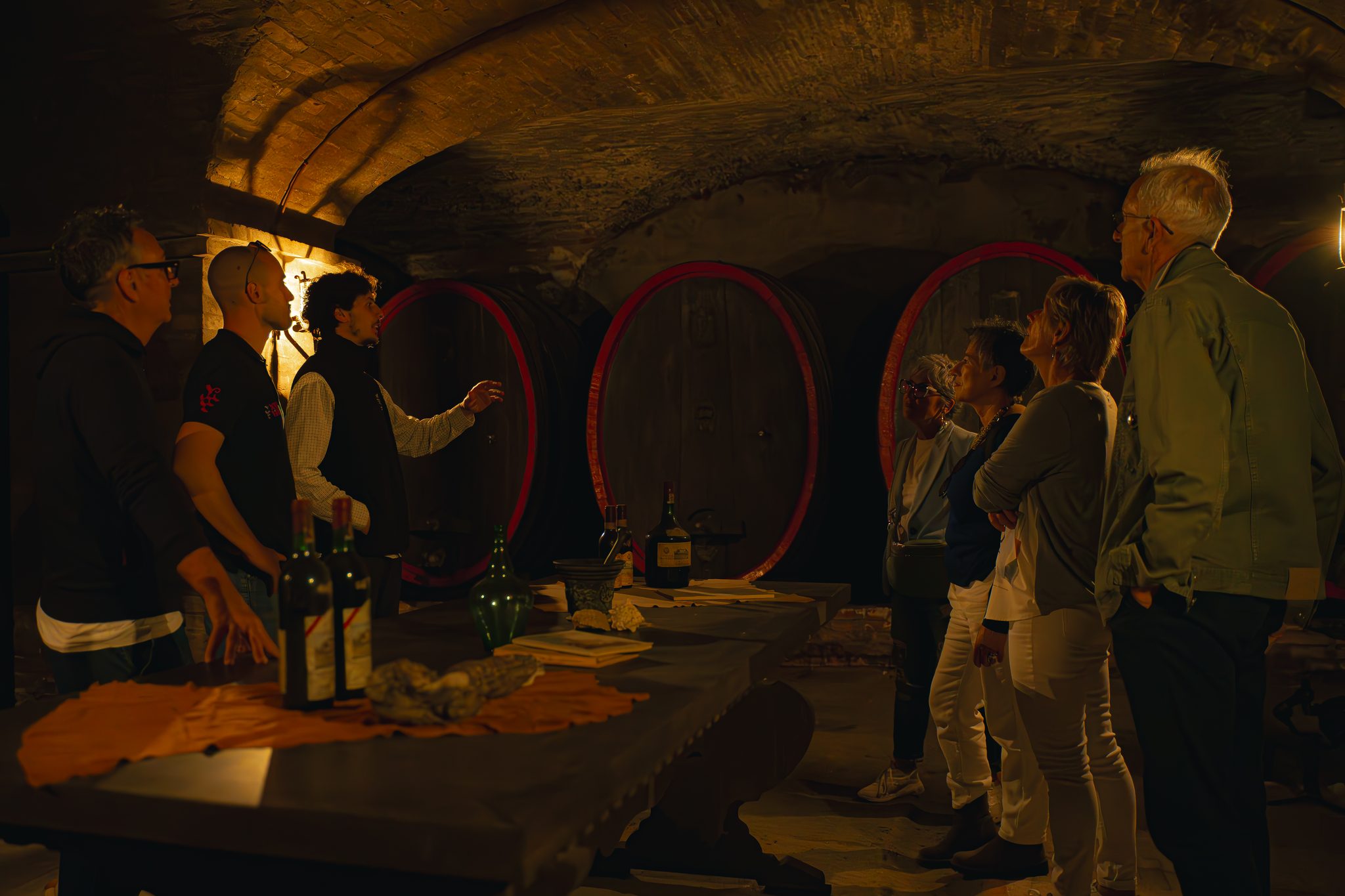 Old wine bottles on a table in a candlelit vaulted cellar, as a small group looks at large wooden casks