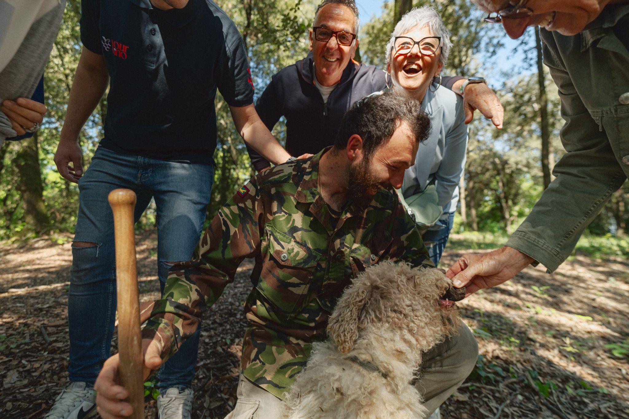 Truffle hunter crouching with his curly white dog as laughing guests gather around a freshly found black truffle in the woods