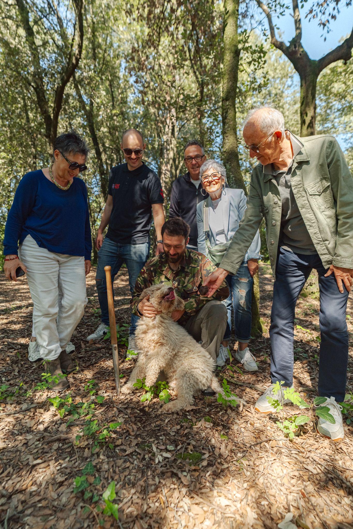 Small group of guests in a sunny wood watching the truffle hunter and his dog next to a truffle spade, one guest holding a black truffle