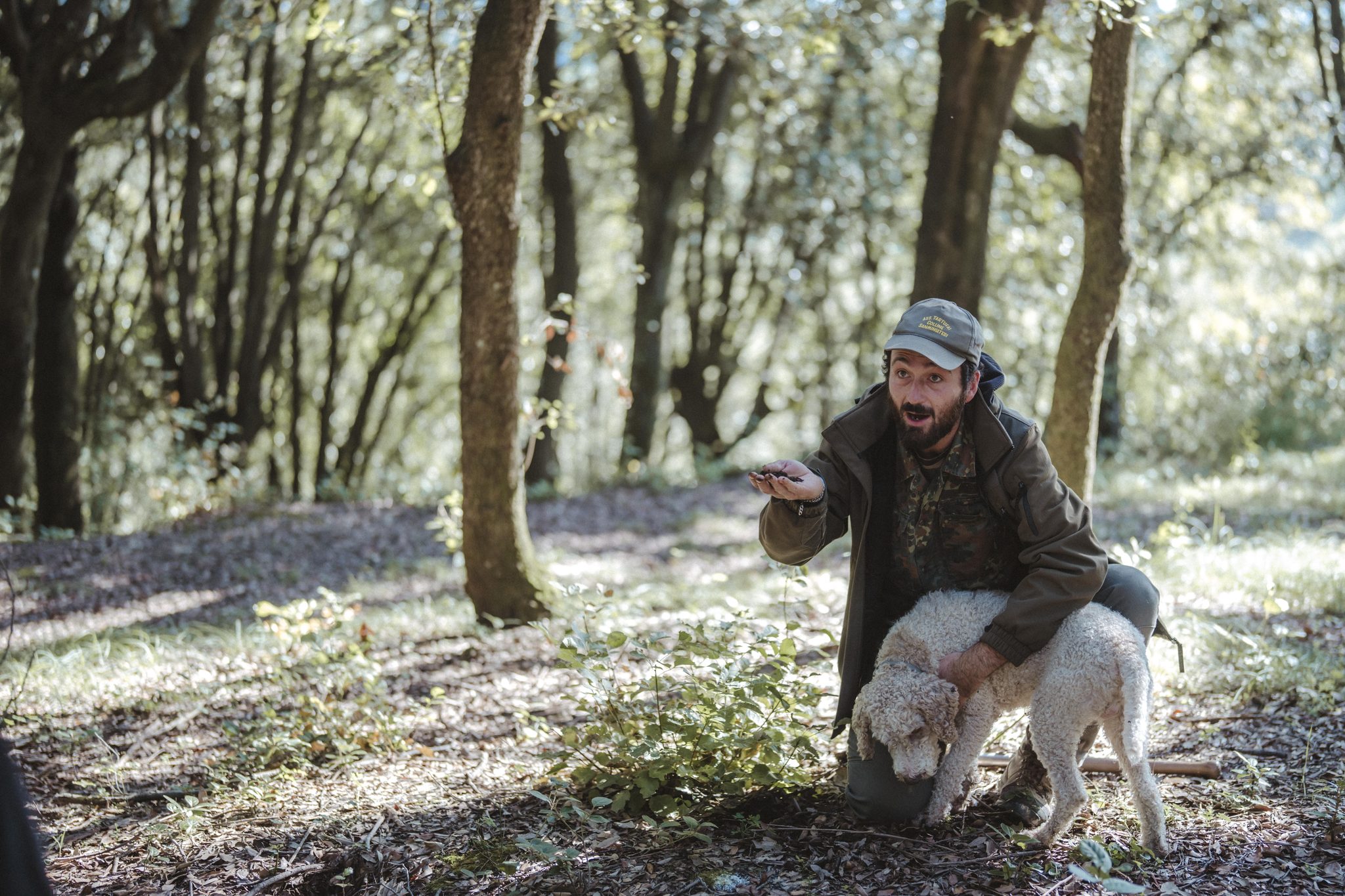 A truffle hunter kneels beside his curly truffle dog in the woods, holding out a handful of soil