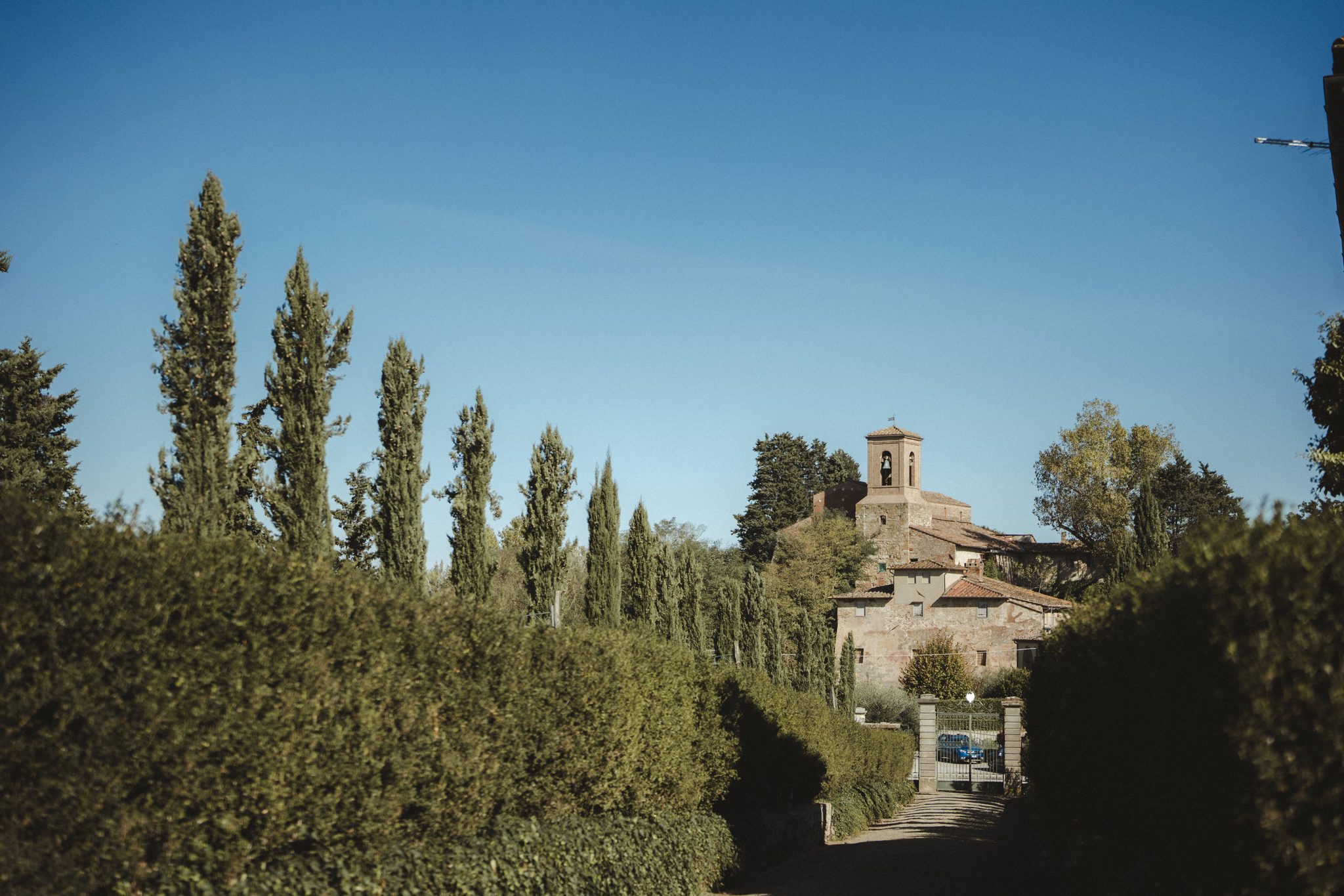 A lane lined with tall cypress trees leads to a small hamlet with a bell tower under a clear blue sky