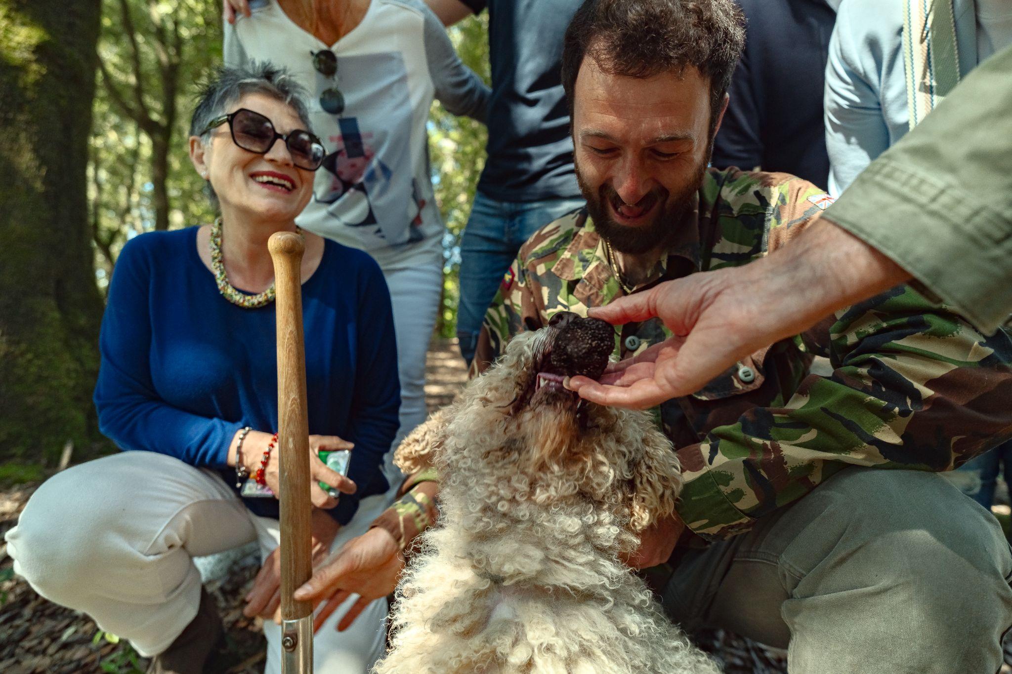 Curly truffle dog sniffing a black truffle held out by a guest while the hunter and a smiling woman look on