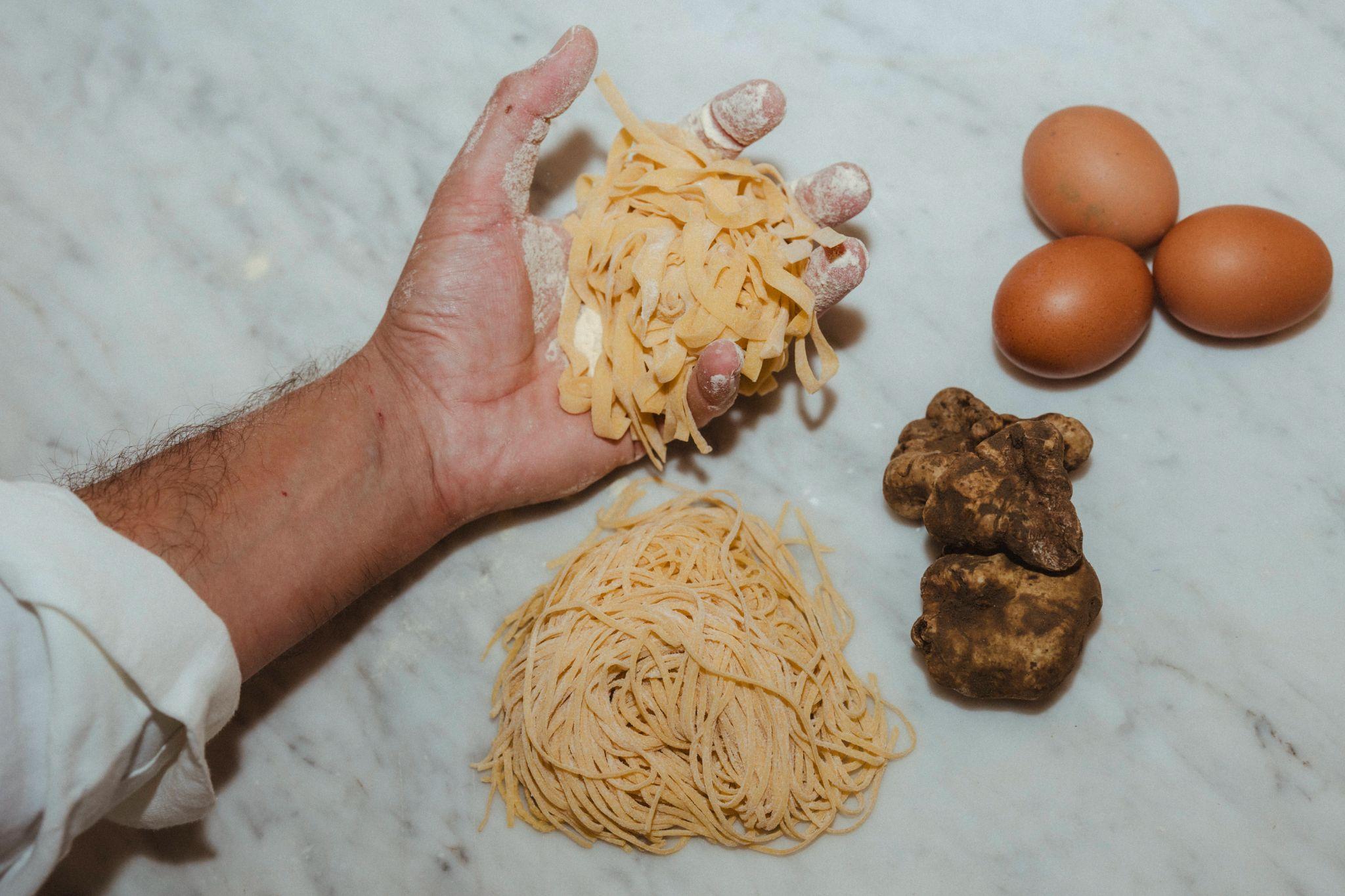 Hand holding fresh tagliatelle above a nest of tagliolini on a marble worktop, with three eggs and pale truffles