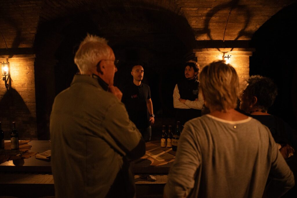 Guests listen to their hosts in a candlelit vaulted wine cellar, with bottles set out on a table