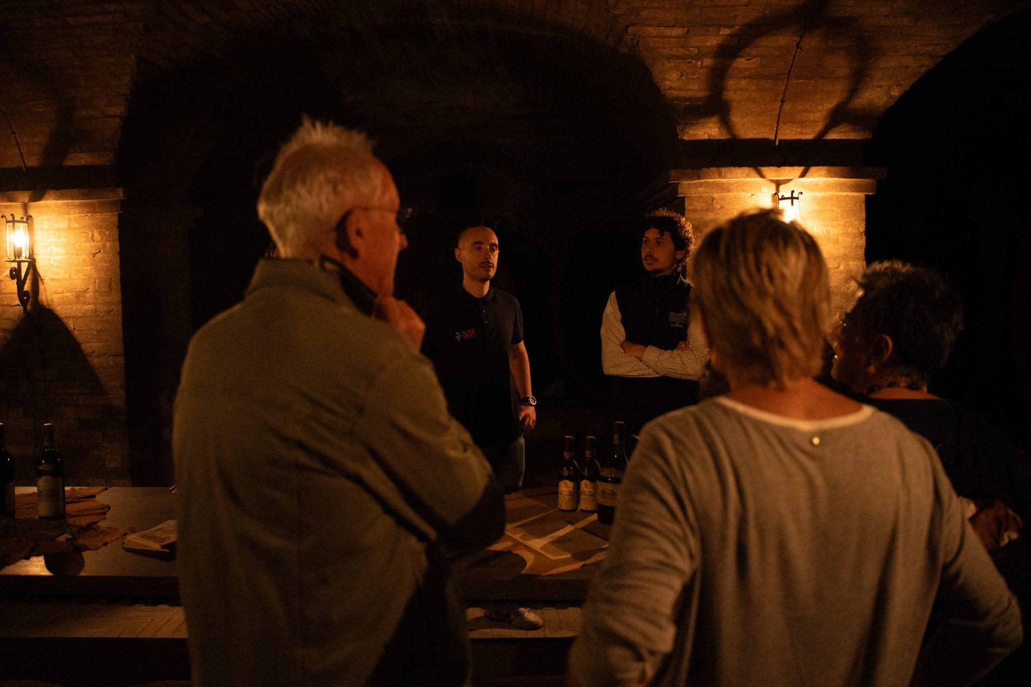 Guests listen to their hosts in a candlelit vaulted wine cellar, with bottles set out on a table