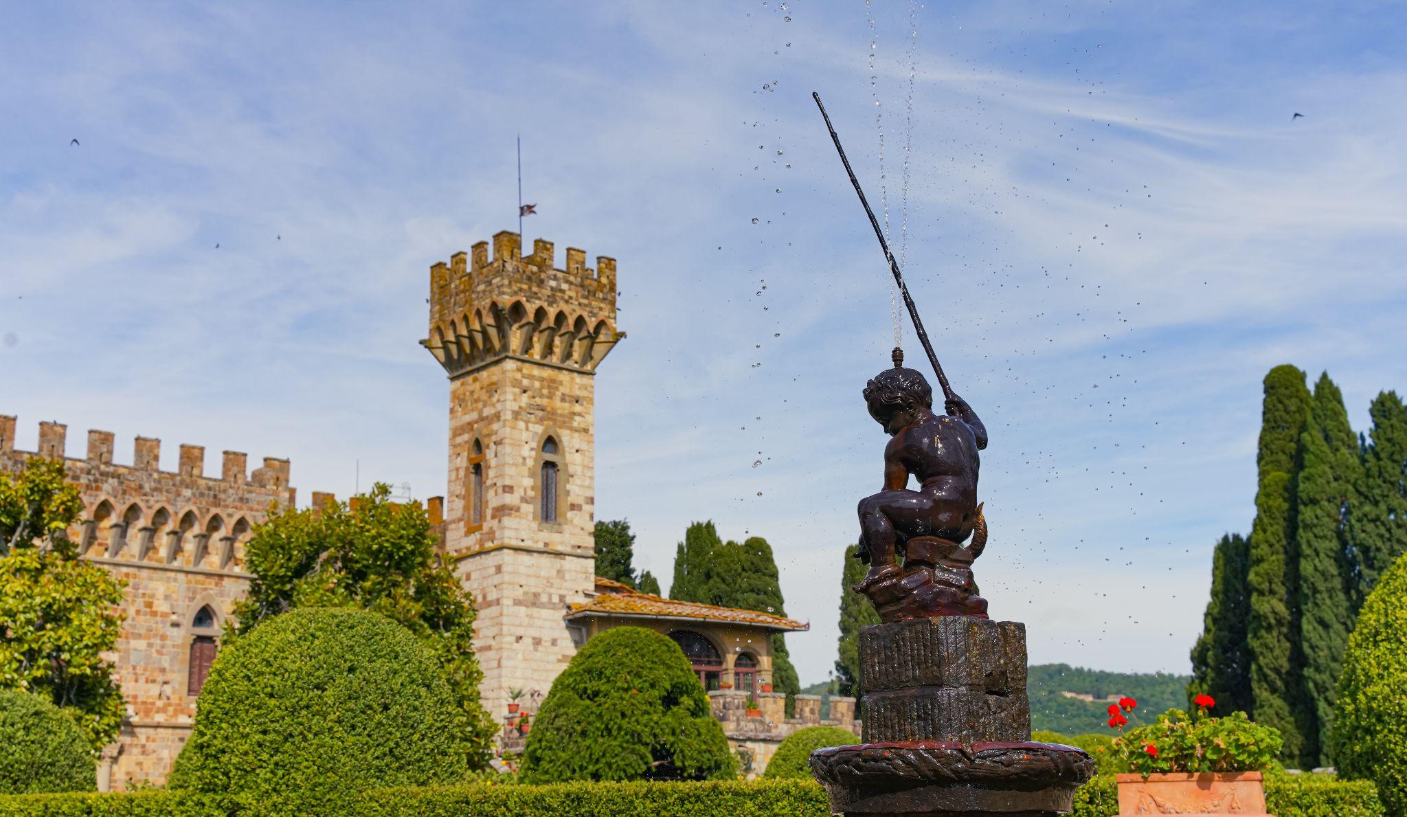 A medieval castle with a crenellated tower behind a garden fountain and tall cypress trees in Tuscany