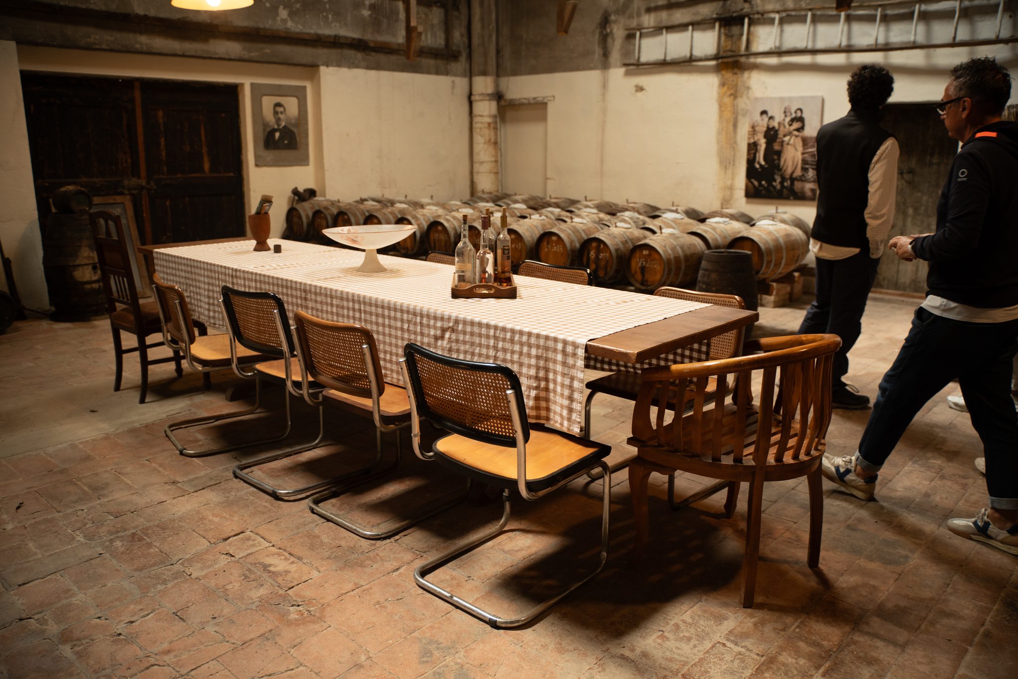 A long table with a checked tablecloth and wine bottles in a historic cellar lined with small wooden barrels