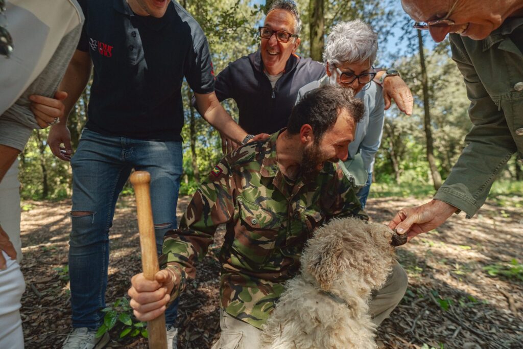 A truffle dog sniffs a freshly found truffle held out by a guest, as the hunter and other guests look on in the woods